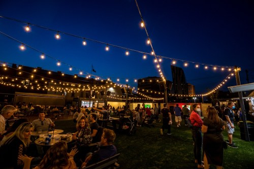 A lively outdoor gathering at night, with string lights illuminating people socializing at tables in a vibrant atmosphere.