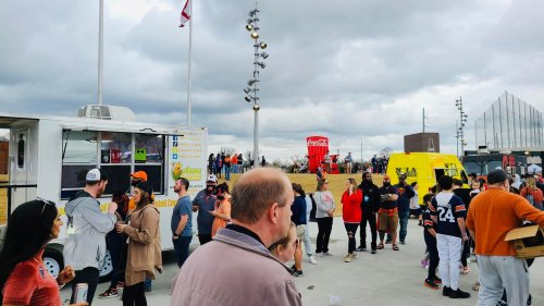 A crowd gathers at a food truck event with flags, a large Coca-Cola sign, and overcast skies in the background.