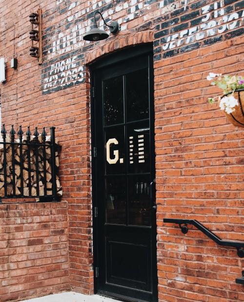 A black door with a large "G" on a brick wall, surrounded by a wrought iron fence and wooden logs stacked nearby.