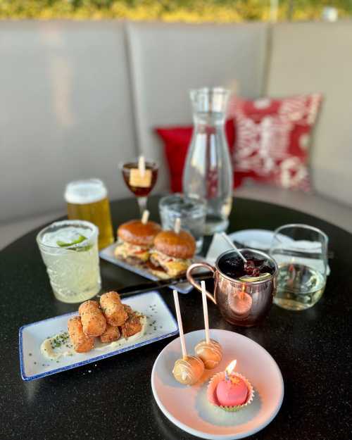 A table with drinks, sliders, and appetizers, including a dessert with a candle, set in a cozy atmosphere.