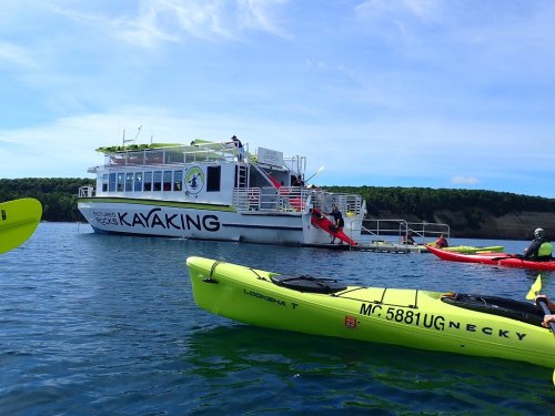 A kayak tour boat is anchored in calm waters, with kayakers paddling nearby against a scenic backdrop.