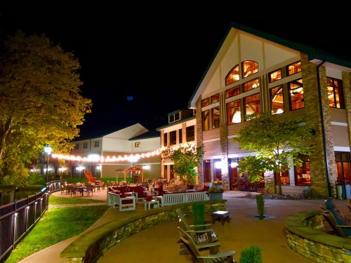 A cozy building at night, illuminated by string lights, surrounded by trees and outdoor seating.