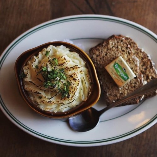 A bowl of creamy mashed potatoes topped with herbs, served with a slice of brown bread and a pat of butter.
