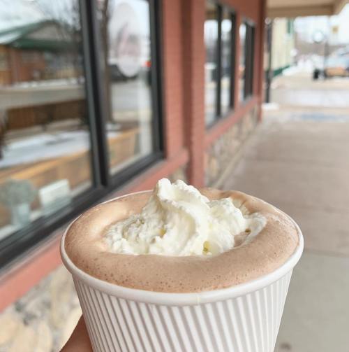 A hand holding a cup of hot chocolate topped with whipped cream, with a storefront visible in the background.