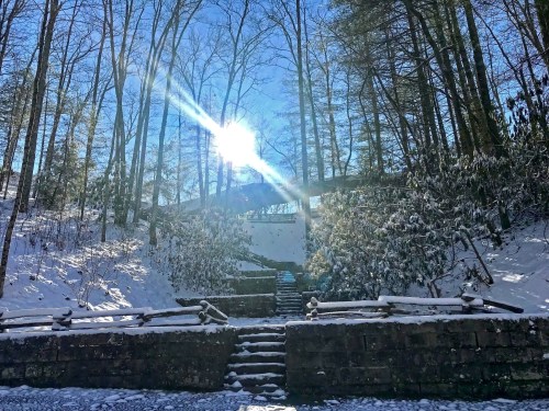 A snowy landscape with steps leading up to a bridge, framed by tall trees and a bright sun shining in the blue sky.