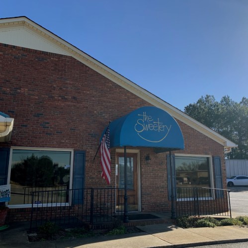 Exterior of a brick building with a blue awning reading "the Sweetery" and an American flag. Clear blue sky above.