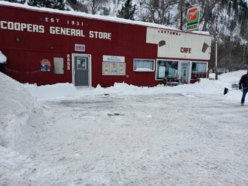 A red general store and café covered in snow, with a snowy parking lot and mountains in the background.