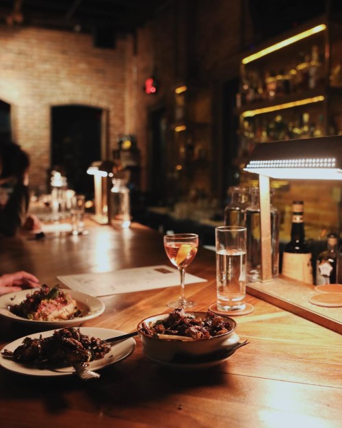 A cozy bar scene with a wooden counter, featuring two plates of food and a cocktail under warm lighting.