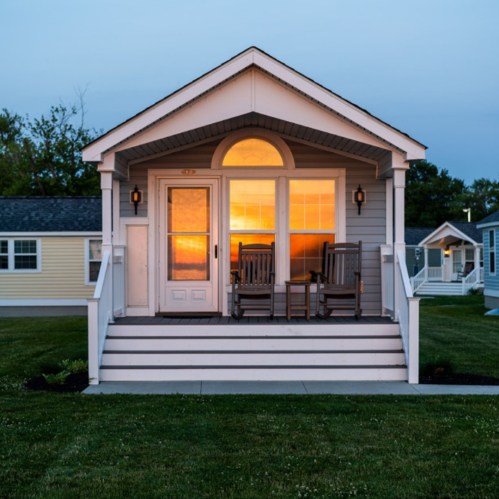 A cozy house with a porch and rocking chairs, illuminated by warm light at sunset. Green lawn in the foreground.
