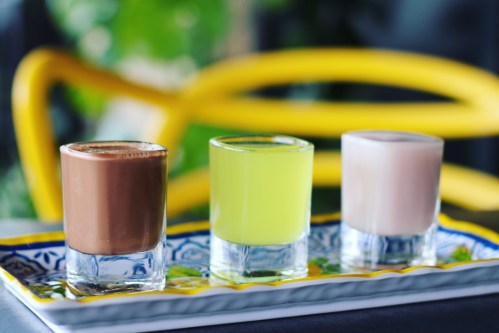 Three shot glasses on a decorative plate: one brown, one green, and one pink, set against a blurred background.