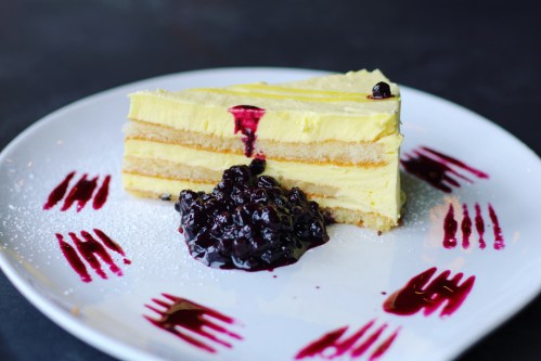 A slice of layered yellow cake topped with berry sauce and served with a dollop of dark berry compote on a white plate.