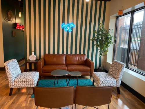 Cozy lounge area with a brown leather sofa, patterned chairs, a coffee table, and a neon sign on striped walls.
