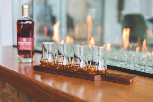 A wooden tray holds four whiskey glasses filled with amber liquid, beside a bottle of whiskey and a glowing fireplace.
