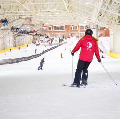 A skier in a red jacket descends a snowy slope indoors, with other skiers and spectators in the background.