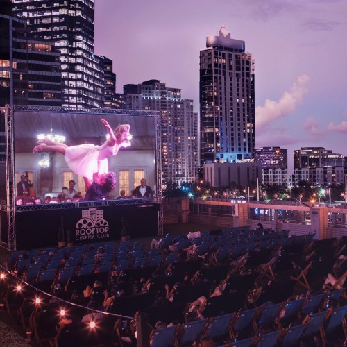 A rooftop cinema with a large screen showing a film, surrounded by an audience and city skyline at dusk.