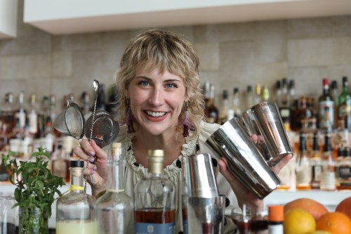 A smiling bartender holds cocktail shakers and strainers, surrounded by various bottles and ingredients on the bar.