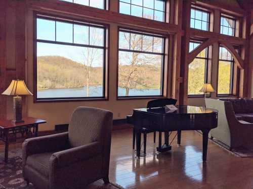 A cozy room with a grand piano, two chairs, and large windows overlooking a serene lake and trees.