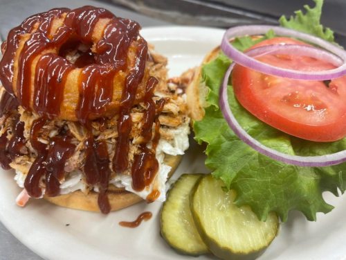 A sandwich with pulled pork, barbecue sauce, a fried onion ring, lettuce, tomato, and pickles on a bun.