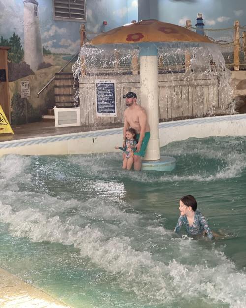 A man and two children play in a wave pool under a colorful mushroom fountain.