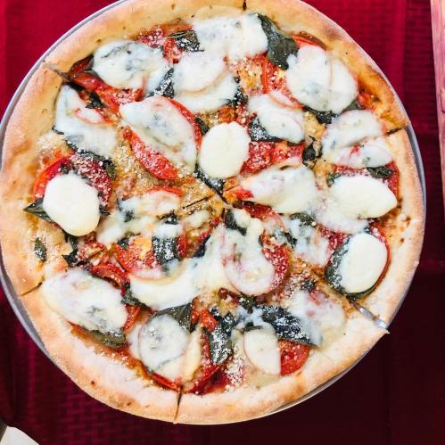 A round pizza topped with tomato slices, fresh basil, mozzarella cheese, and a sprinkle of parmesan on a red tablecloth.