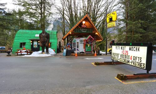 A cozy shop with a green exterior, a large Sasquatch statue, and a sign advertising snacks and gifts in a forested area.