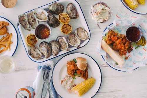 A spread of seafood and fried dishes, including oysters, a sandwich, coleslaw, corn, and fries on a table.