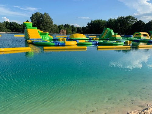 Colorful inflatable water park structures on a clear lake, surrounded by trees and a blue sky.
