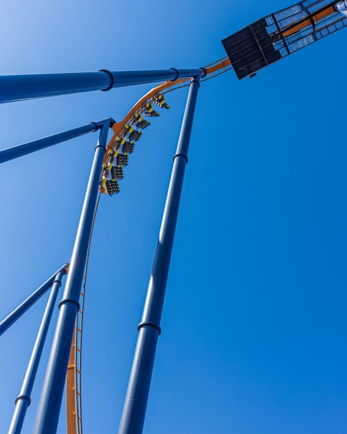 A roller coaster track curves upward against a clear blue sky, with a train of riders approaching the peak.