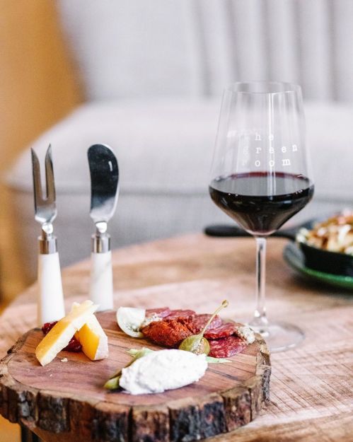 A wooden platter with cheese, meats, and garnishes, alongside a glass of red wine and utensils on a rustic table.
