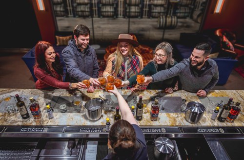 A group of six people toasting with drinks at a bar, surrounded by bottles and a stylish interior.