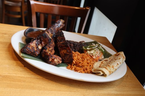A plate of grilled ribs, rice, black beans, salad, and a flatbread, served with a side of sauce.