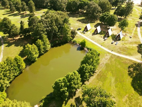 Aerial view of a serene pond surrounded by trees, with three A-frame cabins and a dirt path nearby.