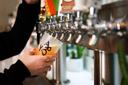 A hand pours a drink from a tap into a clear cup at a bar, with various beer taps in the background.