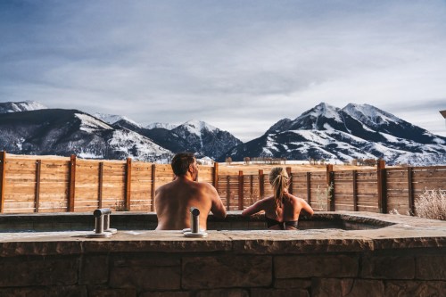 A couple relaxes in a hot tub with snowy mountains in the background under a clear sky.