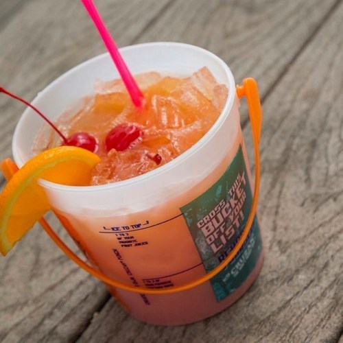 A colorful cocktail in a clear bucket cup, garnished with cherries and an orange slice, resting on a wooden surface.