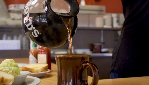 A coffee pot pours hot coffee into a brown mug on a wooden table, with toast and green food in the background.