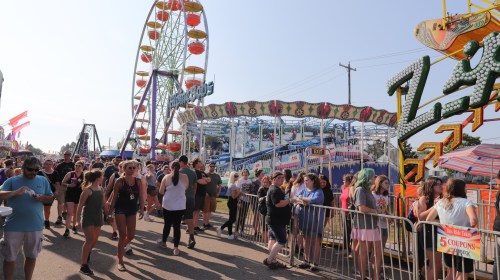 A lively fair scene with people walking, colorful rides, and a Ferris wheel in the background under a clear blue sky.