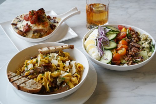 A table setting with three dishes: pasta with meat, a salad with vegetables, and a baked dish, alongside a drink.