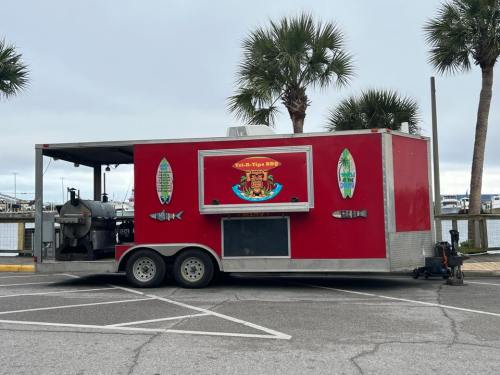 A red food truck with a grill, parked by a marina, surrounded by palm trees and a cloudy sky.
