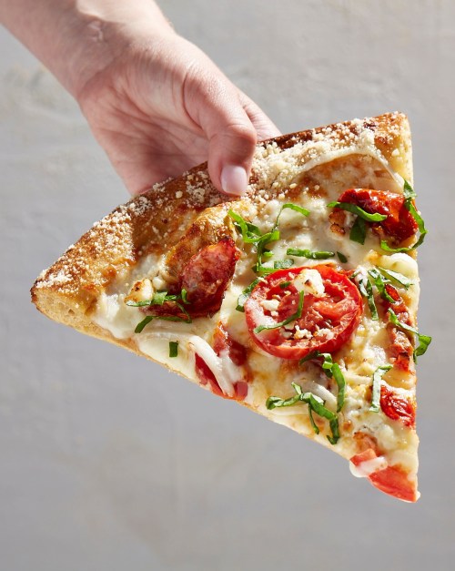 A hand holding a slice of pizza topped with tomatoes, cheese, onions, and fresh basil.