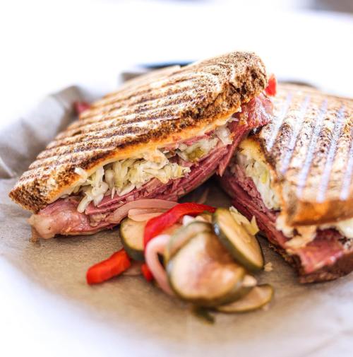 A toasted sandwich filled with corned beef, sauerkraut, and pickles, served with colorful sliced vegetables on the side.