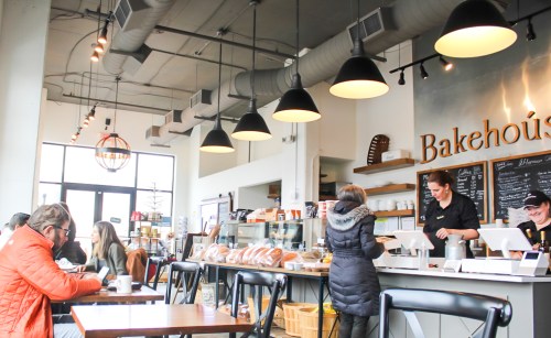 A cozy café interior with customers at tables, a counter with baked goods, and a barista serving at the register.