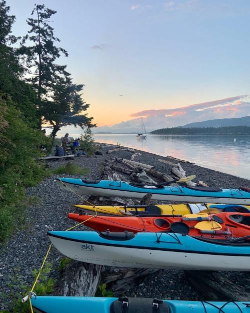 Colorful kayaks lined up on a rocky shore at sunset, with trees and a calm waterway in the background.