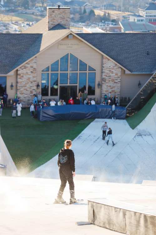 A skier stands on a slope, looking towards a large building with people gathered outside in the background.