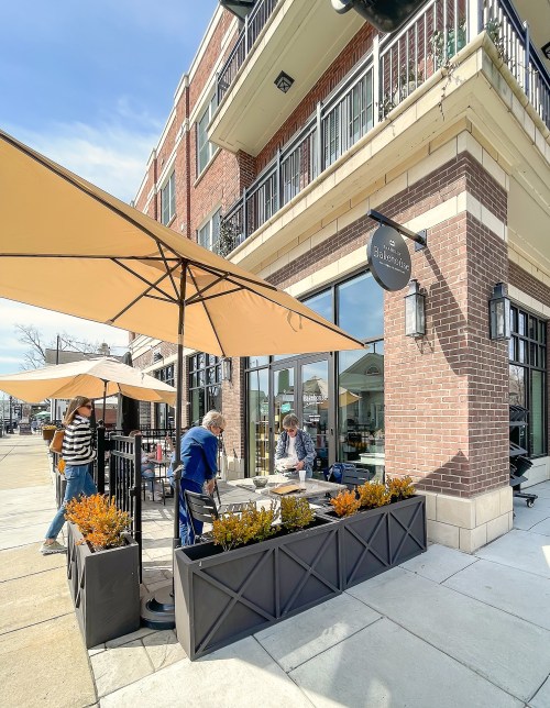 A sunny outdoor café scene with patrons at tables, umbrellas, and planters along a brick building.