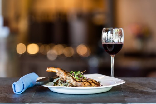A grilled pork chop on a plate with herbs, served alongside a glass of red wine and a blue napkin.
