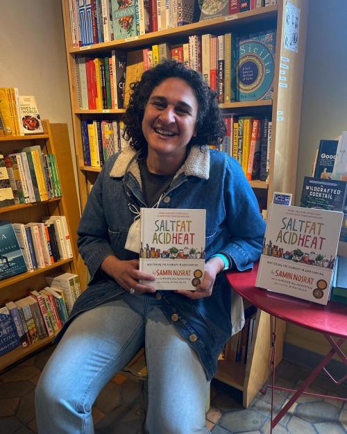 A smiling person holds the book "Salt, Fat, Acid, Heat" in a cozy bookstore filled with colorful books.