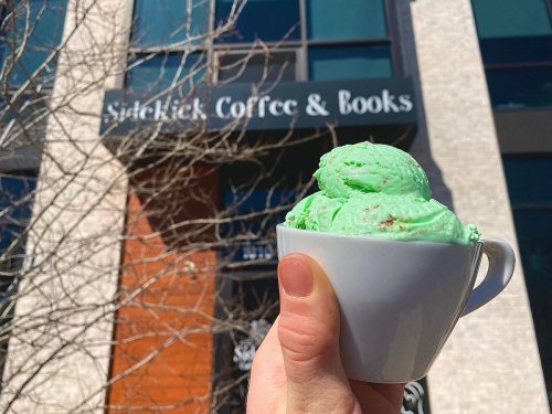 A hand holds a cup of green ice cream in front of Sidekick Coffee & Books.