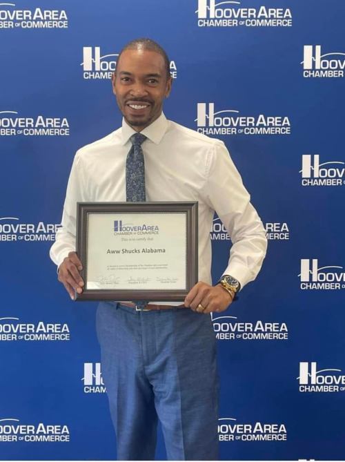 A man in a white shirt and tie holds a certificate in front of a blue backdrop with "Hoover Area Chamber of Commerce."