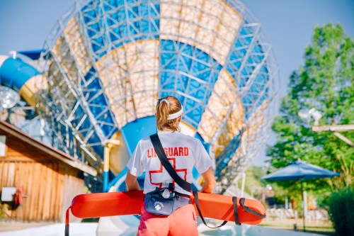 A lifeguard in a red swimsuit stands with a rescue buoy, facing a large water slide in a sunny outdoor setting.